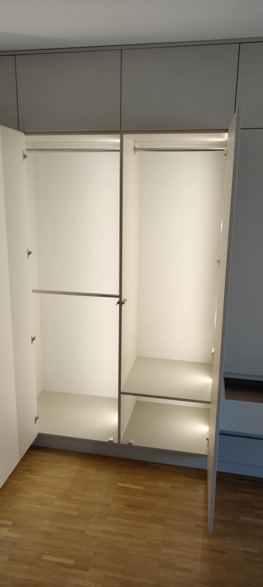 Wnętrze szafy wnękowej z oświetleniem LED, widoczne półki i drążki na ubrania. Jedne drzwi szafy są otwarte, ukazując drewnianą podłogę.