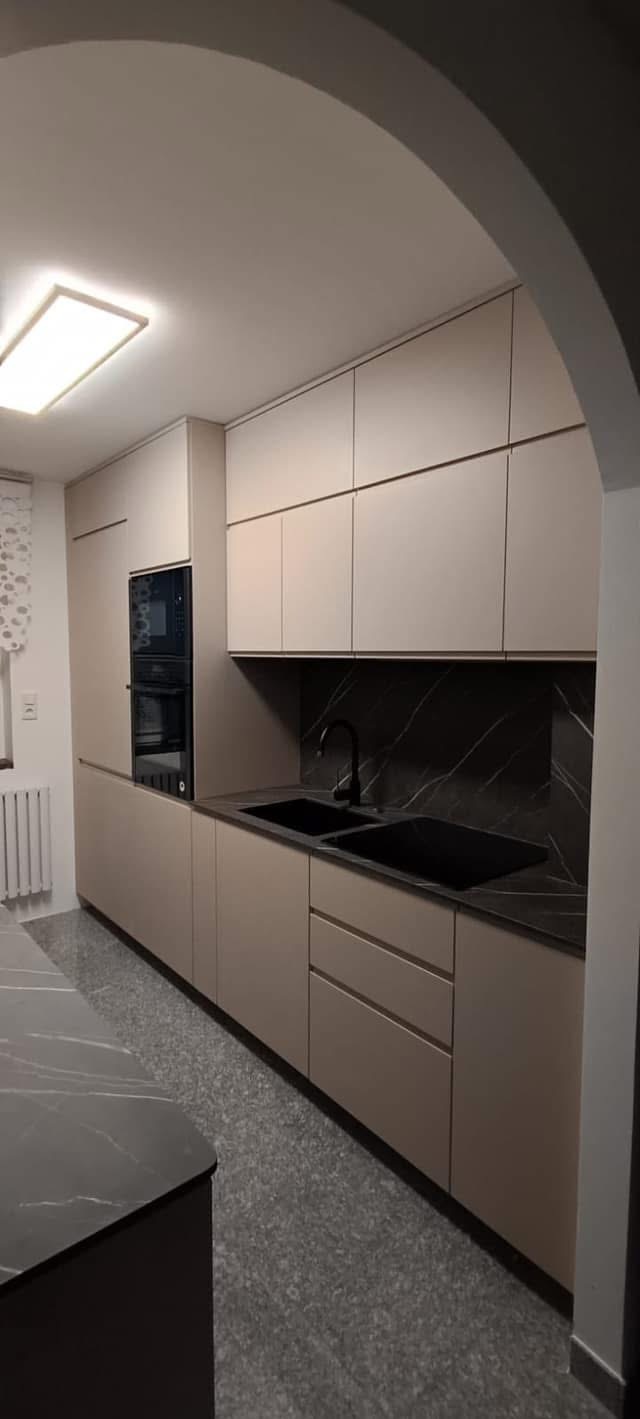 Beżowa kuchnia na wymiar z czarnym blatem i zlewem, oświetlona lampą sufitową. Widok przez łuk architektoniczny.