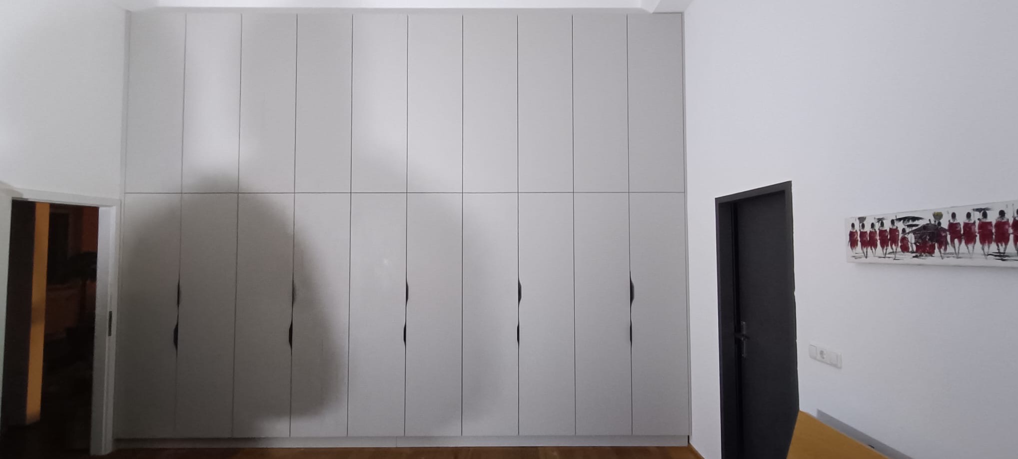 Jasnoszara szafa wnękowa od podłogi do sufitu, z delikatnymi, czarnymi uchwytami, w minimalistycznym wnętrzu z obrazem na ścianie.