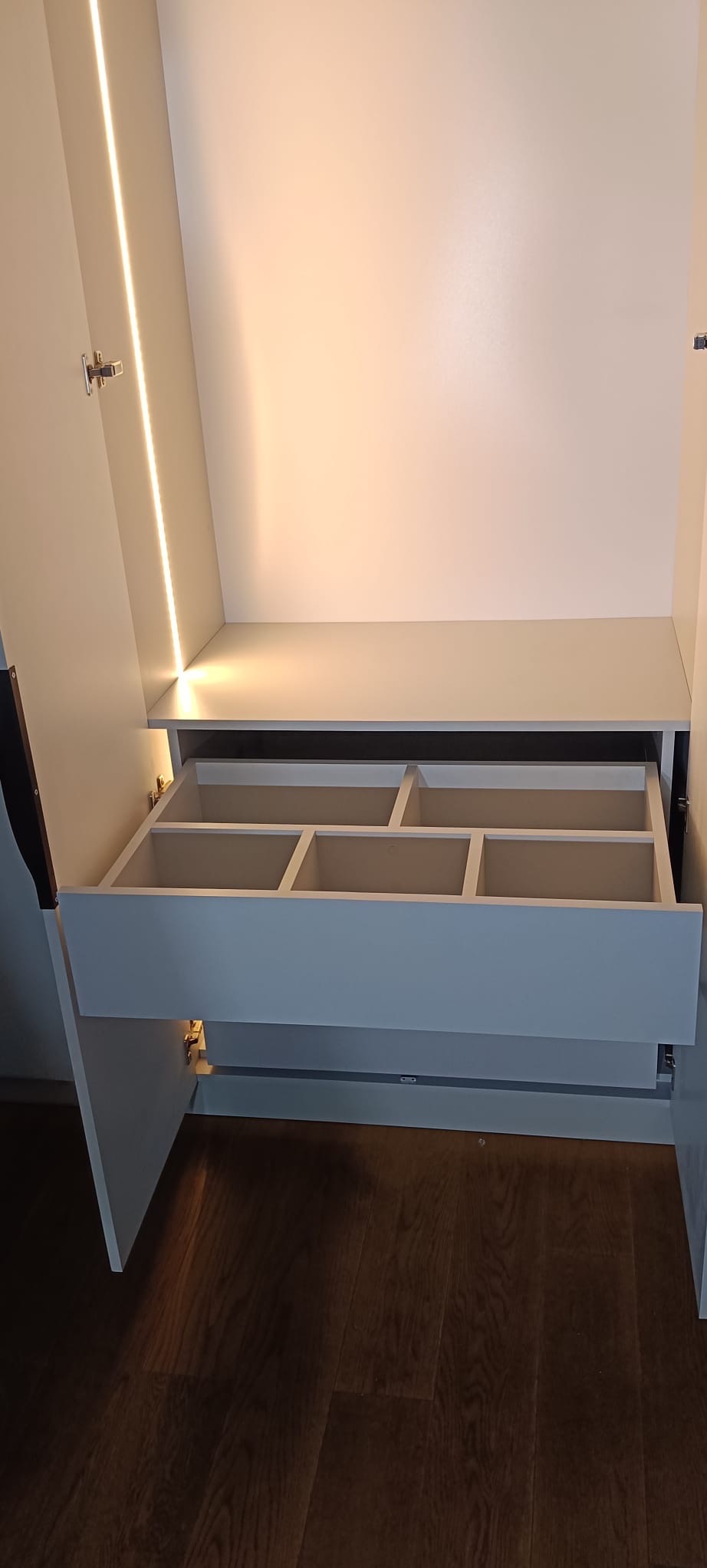 Wnętrze szafy wnękowej z wysuwaną szufladą z przegródkami, oświetloną listwą LED. Widoczne zawiasy i drewniana podłoga. Jasne kolory, minimalistyczny design.