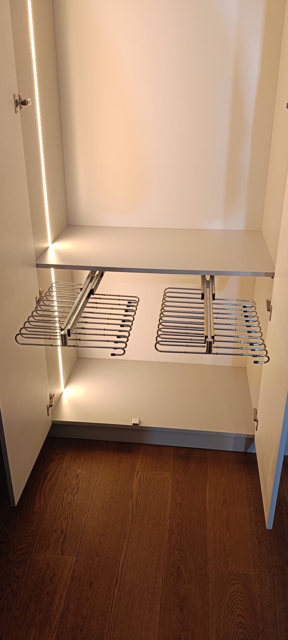Wnętrze szafy wnękowej z wysuwanymi wieszakami na spodnie, oświetlone listwami LED. Widoczna drewniana podłoga i białe półki.
