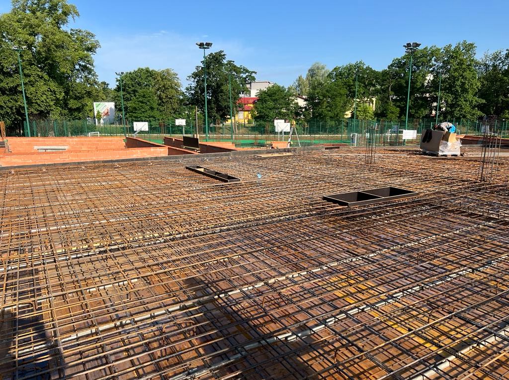 Siatka zbrojeniowa na budowie, widok z góry, z elementami szalunku i ceglanym murem w tle, w słoneczny dzień. Detaliczna struktura zbrojenia.