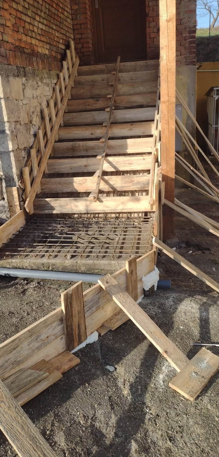 Wykonywanie betonowych schodów zewnętrznych z drewnianym szalunkiem i zbrojeniem. Widoczna murowana ściana budynku w tle.