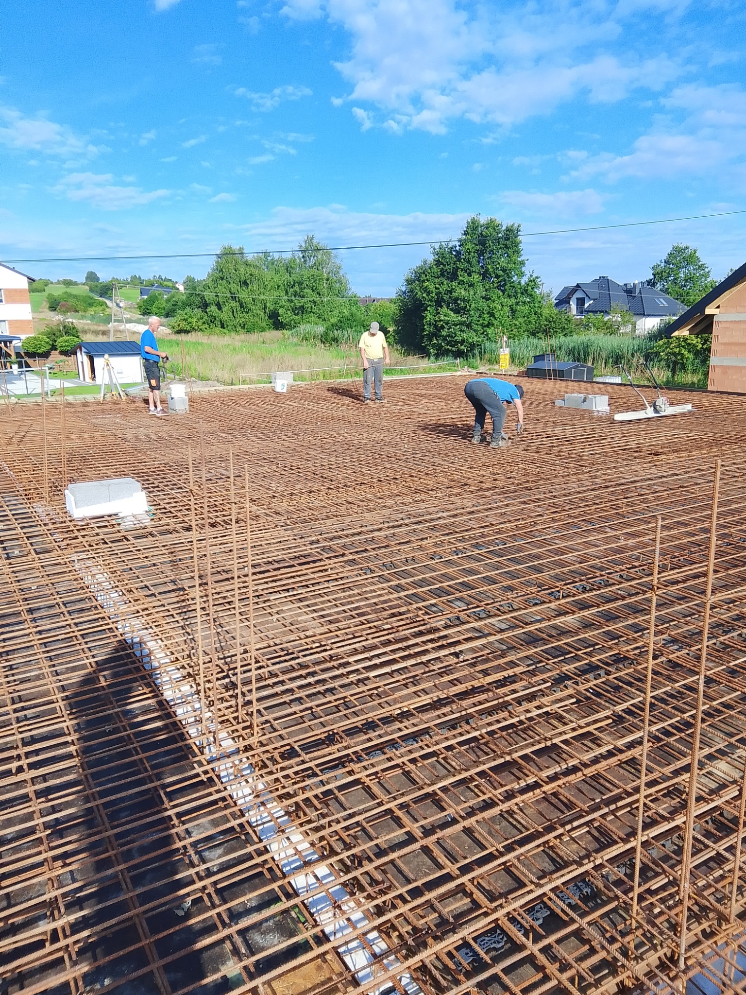 Zbrojenie fundamentów domu jednorodzinnego z widocznymi pracownikami budowlanymi na tle zieleni i błękitnego nieba. Widoczne elementy szalunku i narzędzia.