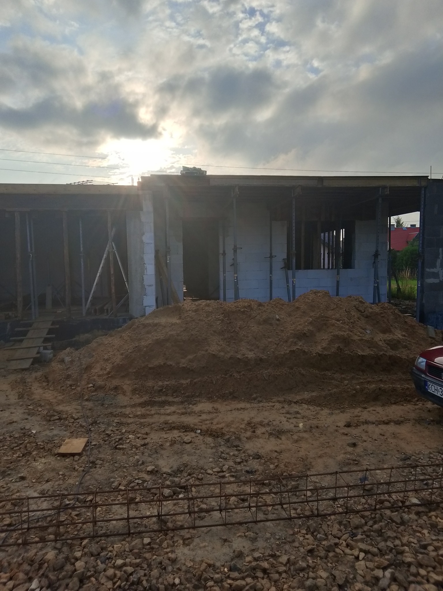 Stan surowy otwarty domu z białych bloczków, widoczne zbrojenia i sterta ziemi. Na pierwszym planie kamienie i zbrojenie. Słoneczny dzień, częściowe zachmurzenie.