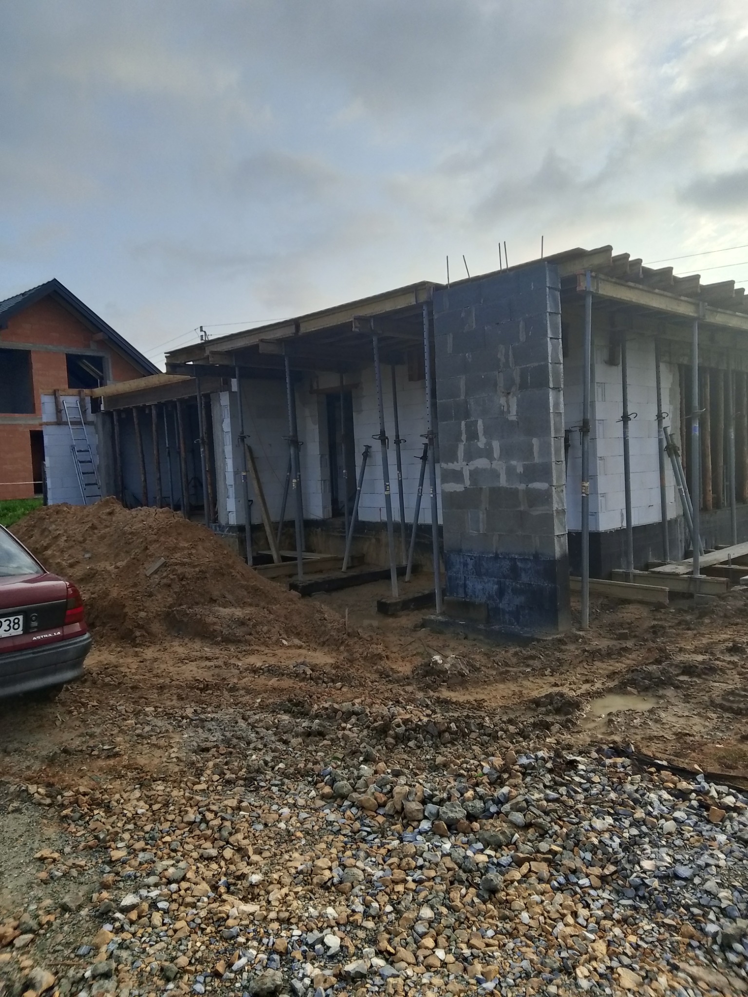 Budowa domu w trakcie: ściany z bloczków, podpory stropu, widoczny fundament izolowany. W tle drugi budynek w stanie surowym otwartym i samochód osobowy.