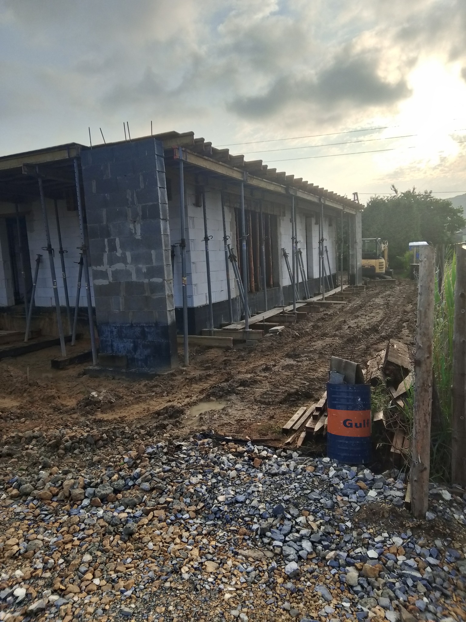 Budowa domu w trakcie: ściany z bloczków, podpory, widoczny sprzęt budowlany w tle. Ujęcie z poziomu gruntu, niebo pochmurne.