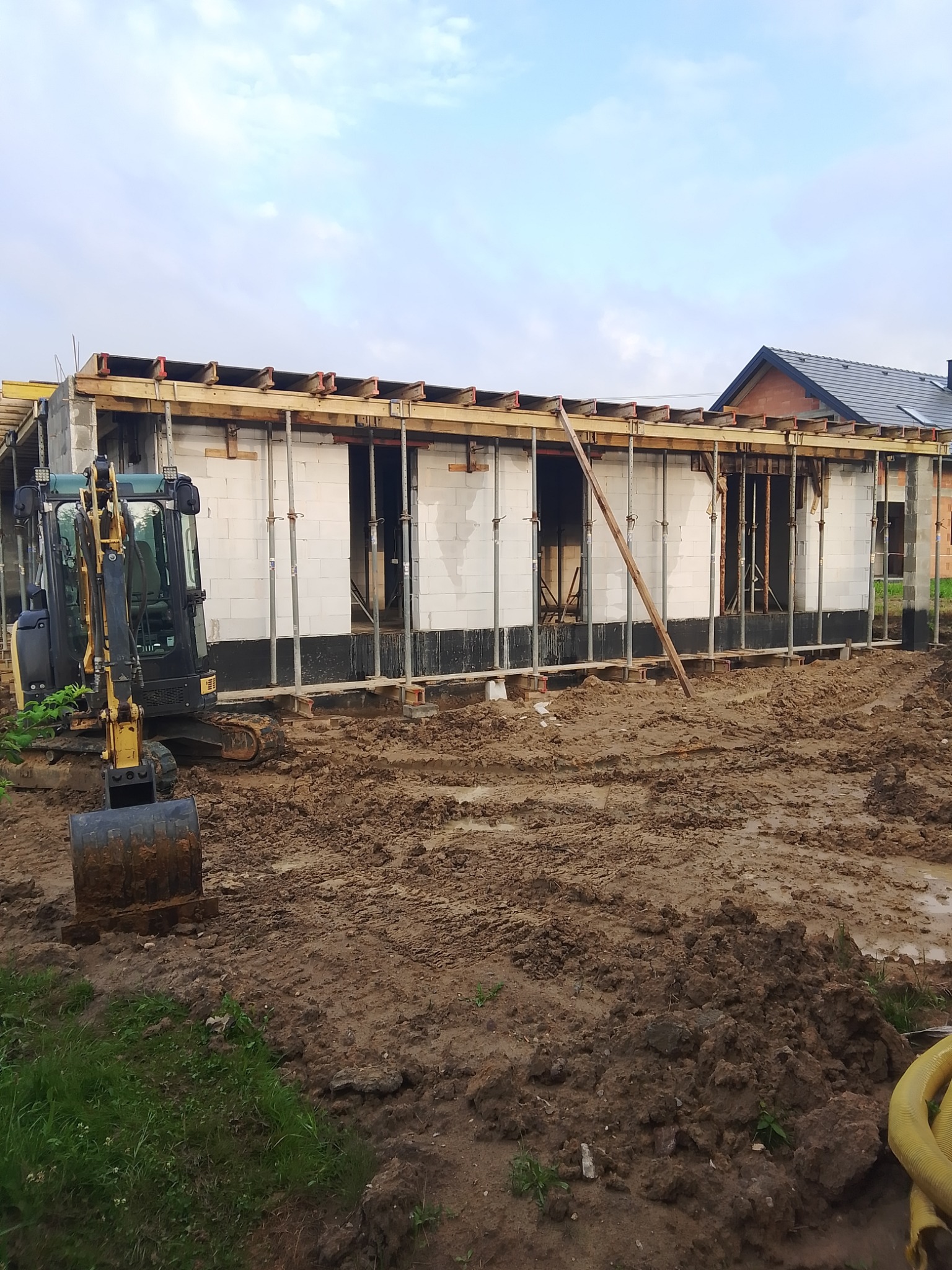Budowa domu z betonu komórkowego: ściany wzniesione, widoczne szalunki, mini koparka na błotnistym placu budowy. Widok na konstrukcję w trakcie realizacji.