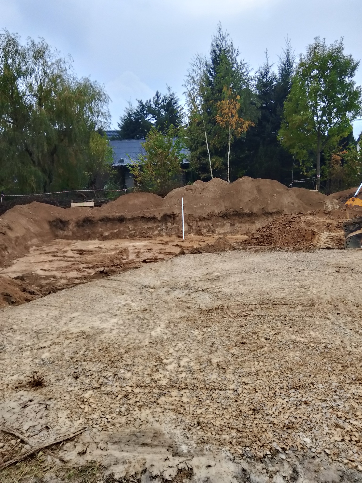 Wykop pod fundamenty domu jednorodzinnego, widoczny z gruntu. W tle drzewa i fragment dachu domu. Słup geodezyjny w wykopie.