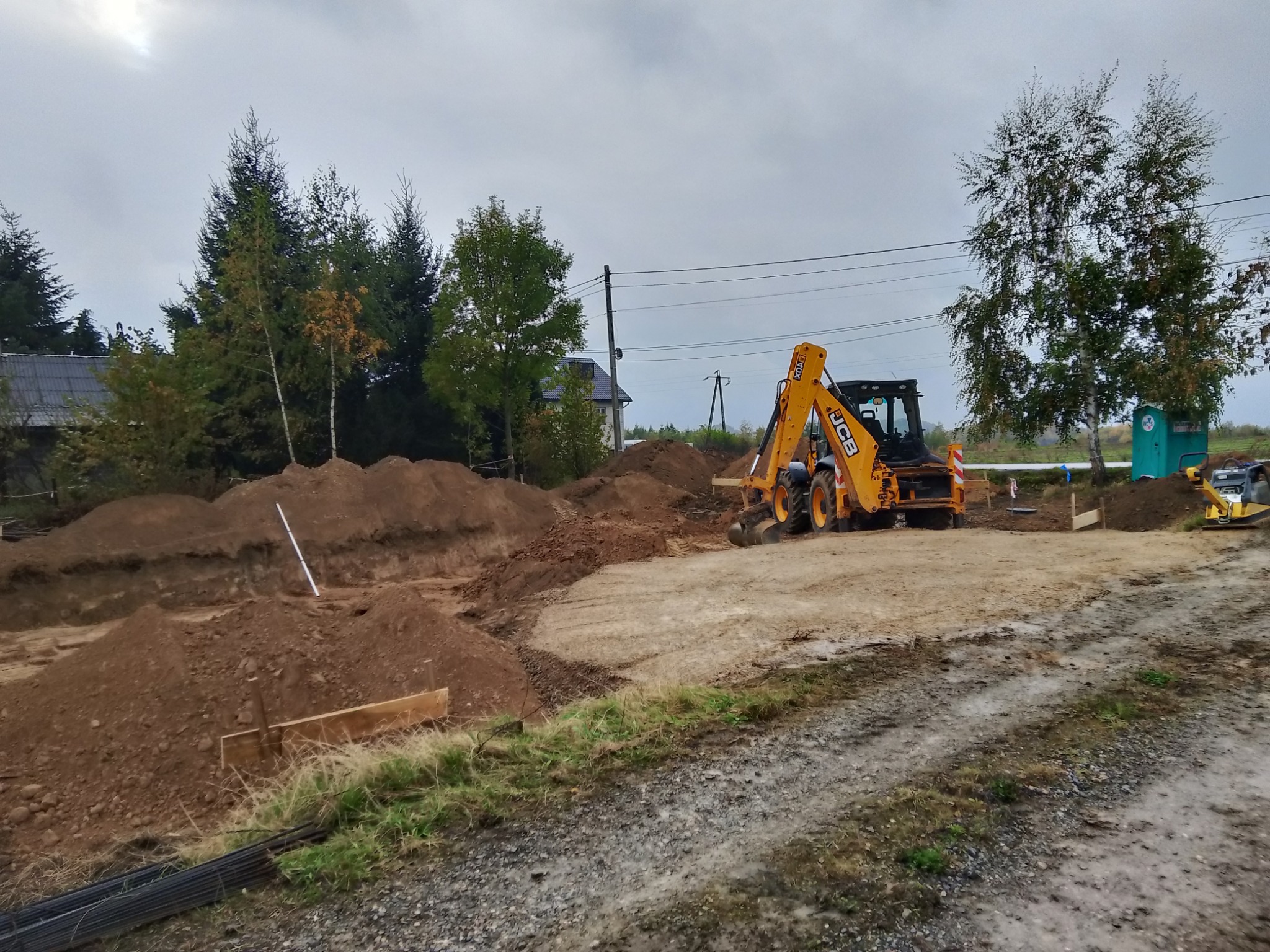 Żółta koparko-ładowarka JCB przy wykopie fundamentów. Na pierwszym planie ziemia, w tle drzewa i budynek. Pochmurne niebo.