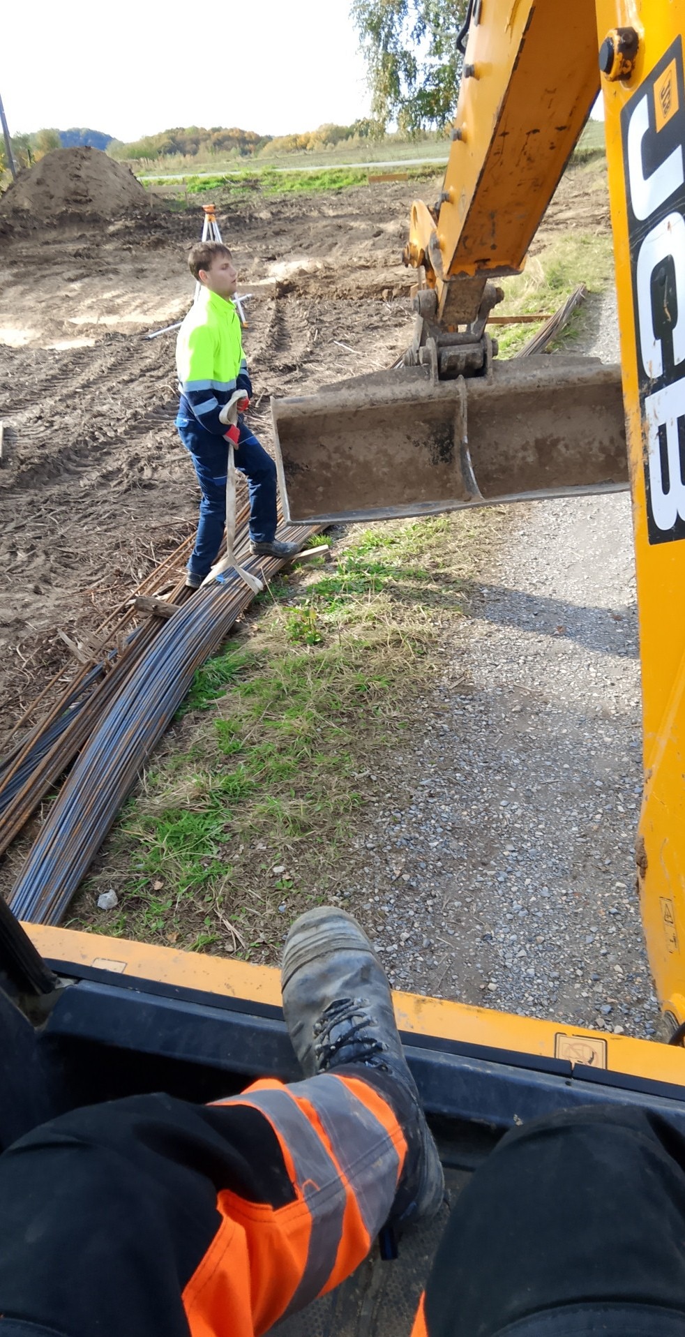 Praca ziemne z użyciem koparki JCB, widok z perspektywy operatora. W tle pracownik w odblaskowej odzieży, stosy zbrojenia i teren budowy.