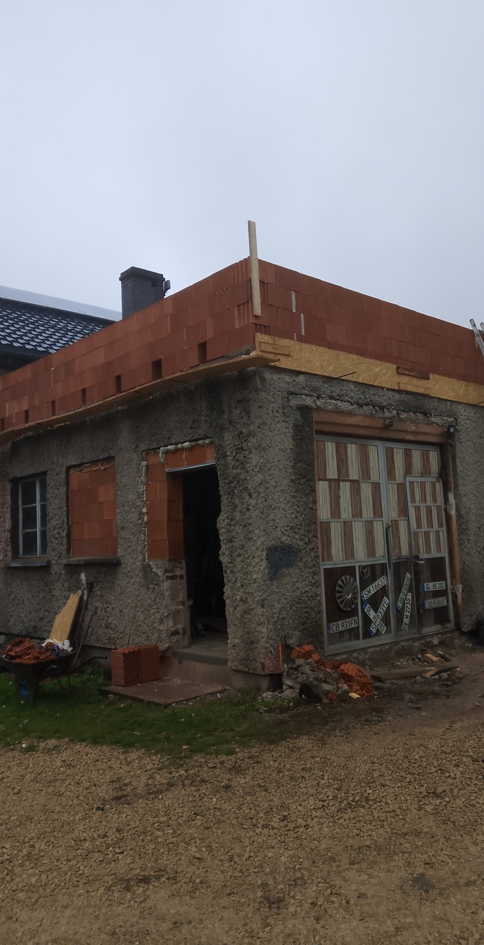 Budynek w trakcie rozbudowy: murowane ściany z czerwonej cegły, surowy beton, garaż z nietypowymi drzwiami i gruz. Widoczny wózek z cegłami.