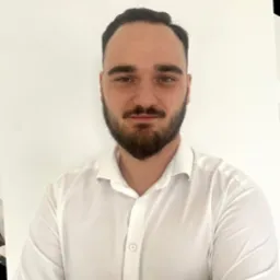 Portret brodatego mężczyzny w białej koszuli na jasnym tle. Kadr obejmuje górną część ciała, z lekkim uśmiechem i bezpośrednim spojrzeniem w obiektyw.