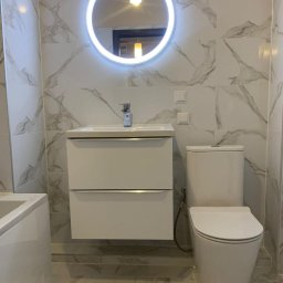 Dav-Bau Dawid Górecki - Biała łazienka z marmurowymi płytkami, okrągłym lustrem z podświetleniem LED, umywalką z szafką i toaletą. Minimalistyczny design, jasne oświetlenie.