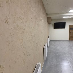 Dav-Bau Dawid Górecki - Beżowa ściana z dekoracyjną fakturą, grzejniki, szara podłoga z płytek, telewizor na ścianie w tle. Widok z boku, oświetlenie sufitowe.