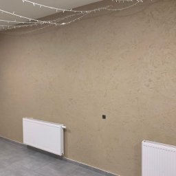 Dav-Bau Dawid Górecki - Beżowa ściana z dekoracyjną fakturą, oświetlona girlandą świetlną. Widoczne dwa białe grzejniki i fragment ściany wykończonej kamieniem dekoracyjnym.