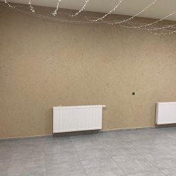 Dav-Bau Dawid Górecki - Beżowa ściana z dekoracyjną fakturą, dwa białe grzejniki, szara podłoga z płytek i girlanda świetlna pod sufitem. Fragment ściany wykończony kamieniem dekoracyjnym.