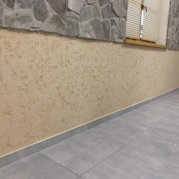 Dav-Bau Dawid Górecki - Ściana z dekoracyjną, beżową strukturą tynku, szara podłoga z płytek, kamienna okładzina ściany i drewniana rama okienna z żaluzjami w jasnym wnętrzu.