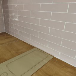 Dav-Bau Dawid Górecki - Ściana wyłożona podłużnymi, beżowymi płytkami z białymi fugami, przylegająca do drewnianego blatu z płytą indukcyjną. Widoczne niedociągnięcia w fugowaniu.