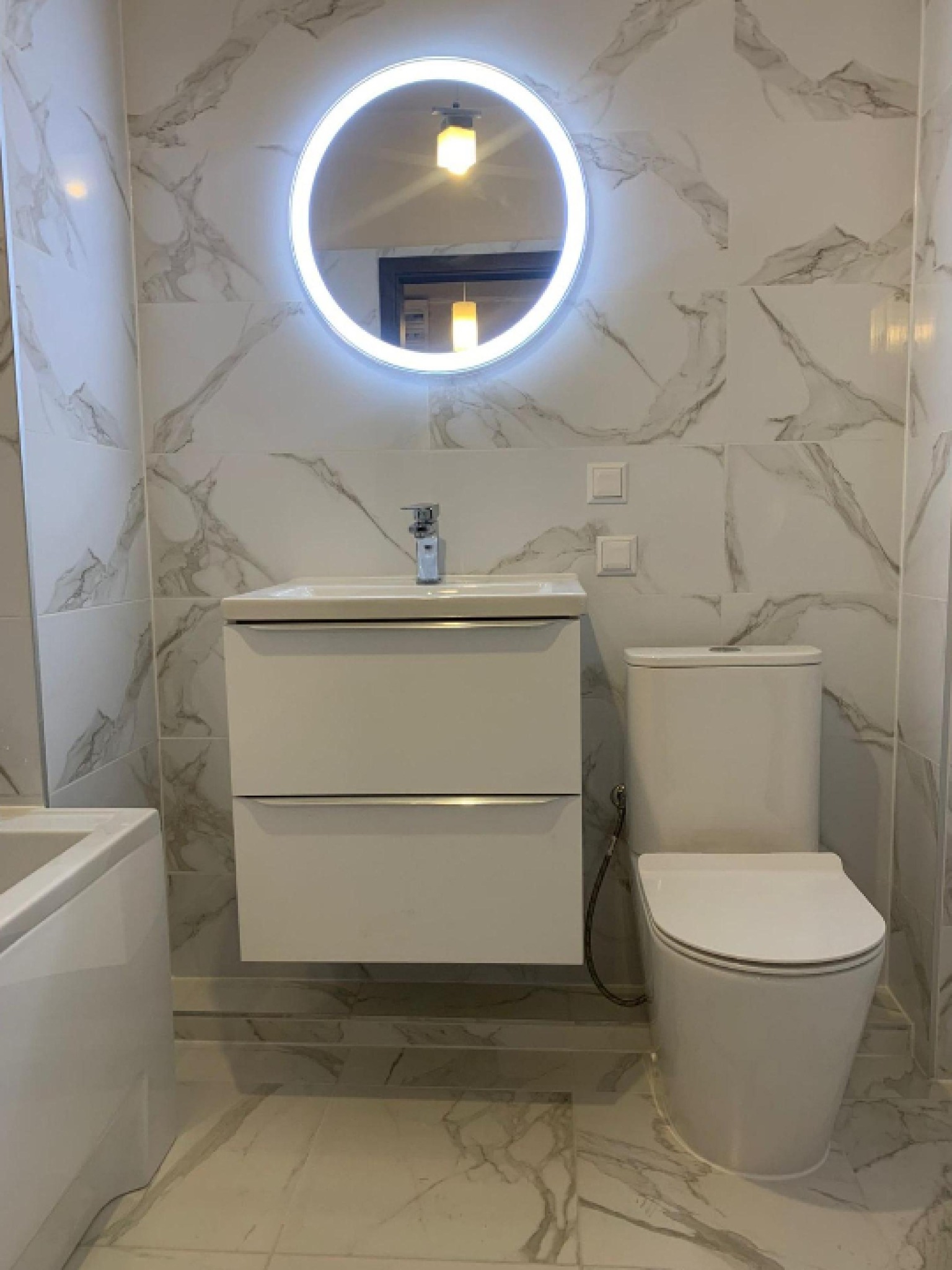 Biała łazienka z marmurowymi płytkami, okrągłym lustrem z podświetleniem LED, umywalką z szafką i toaletą. Minimalistyczny design, jasne oświetlenie.