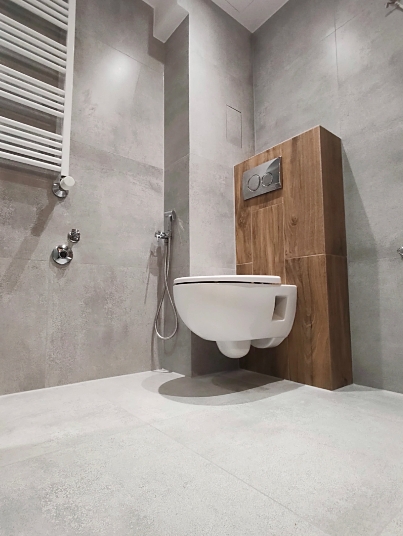 Nowoczesna łazienka z podwieszaną toaletą na drewnianej obudowie, szara płytka imitująca beton, chromowana armatura i grzejnik drabinkowy.