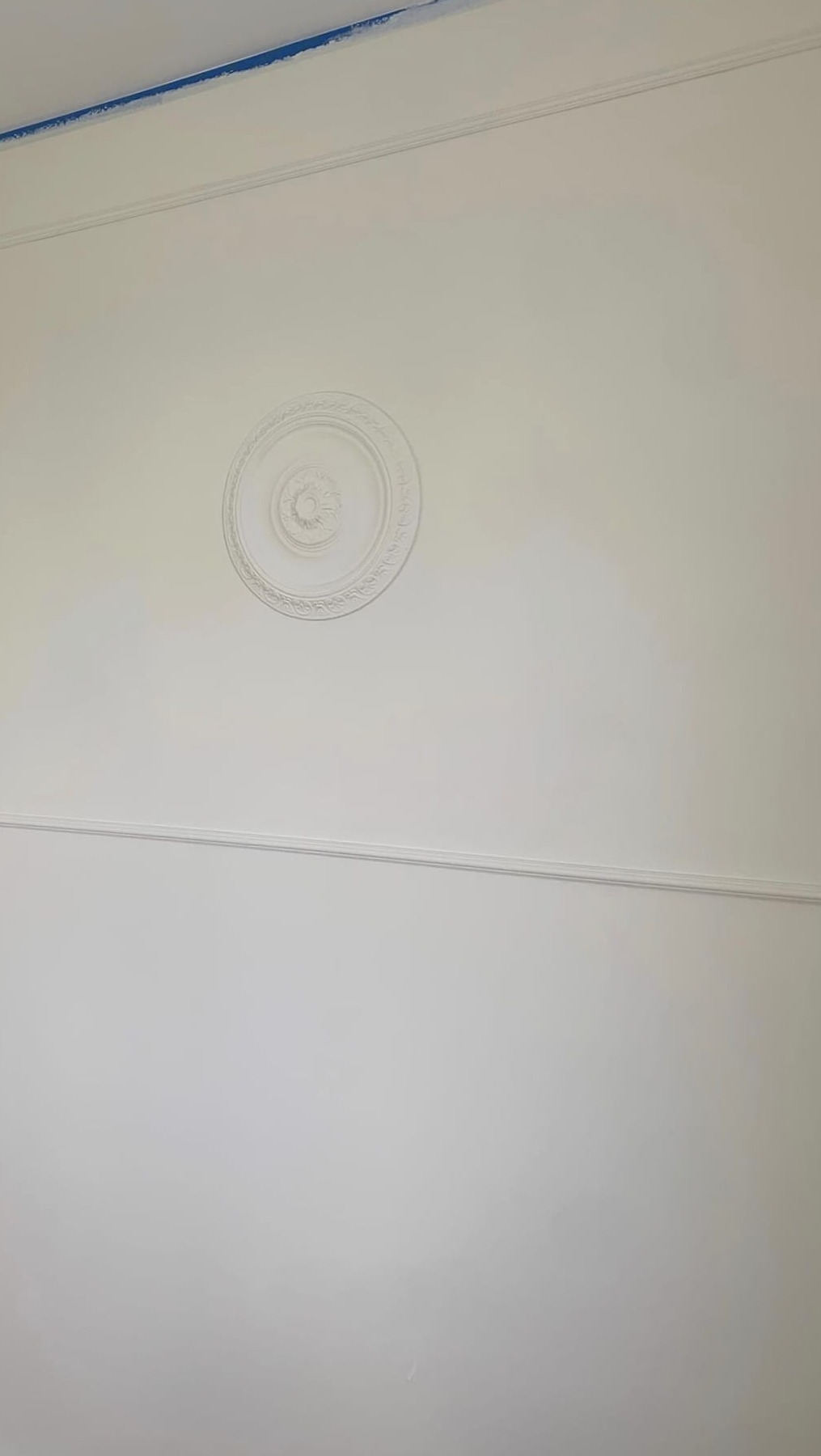 Biała ściana z dekoracyjnym, okrągłym elementem sztukaterii pod sufitem i listwą przypodłogową. Górna krawędź ściany zabezpieczona niebieską taśmą malarską.