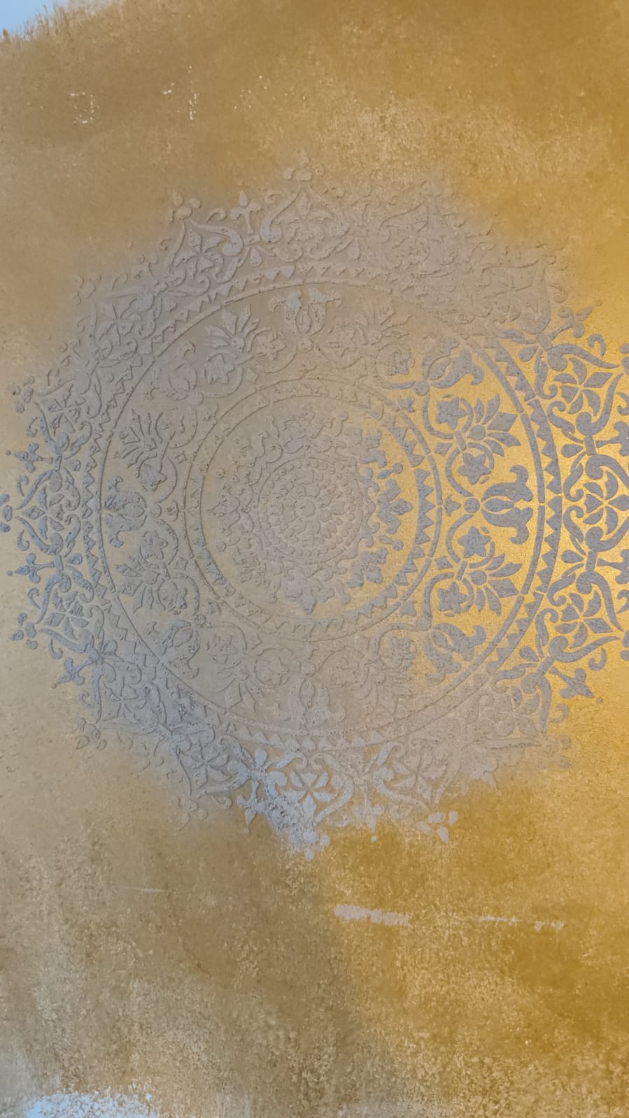 Dekoracyjna ściana z efektem stiuku w odcieniach złota i szarości, z wyraźnym, symetrycznym wzorem mandali. Unikalna faktura i gra światła podkreślają artystyczny charakter.