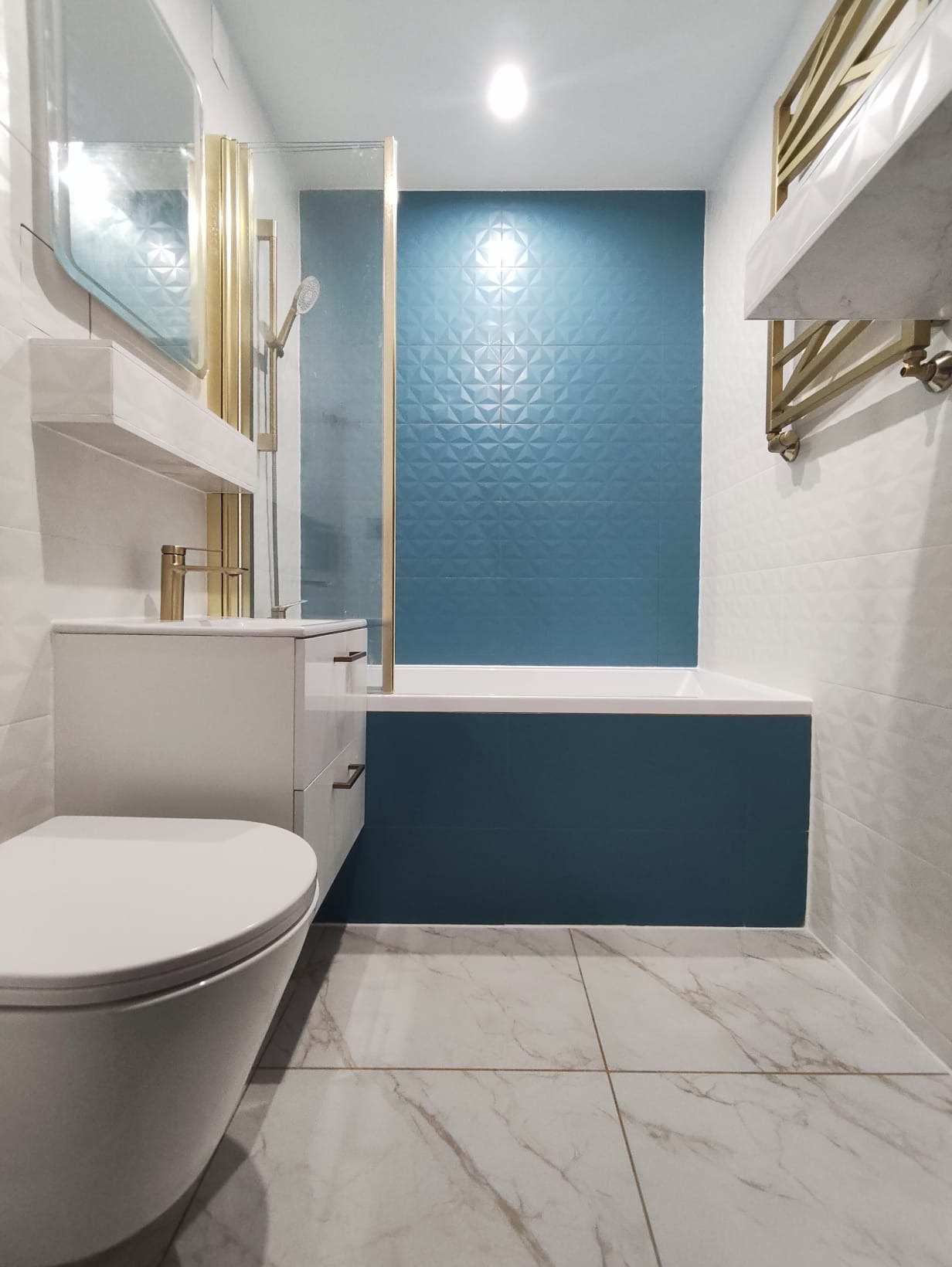 Nowoczesna łazienka po remoncie: biało-turkusowe płytki z geometrycznym wzorem, złota armatura, wisząca szafka z umywalką i toaleta. Marmurowe płytki podłogowe.