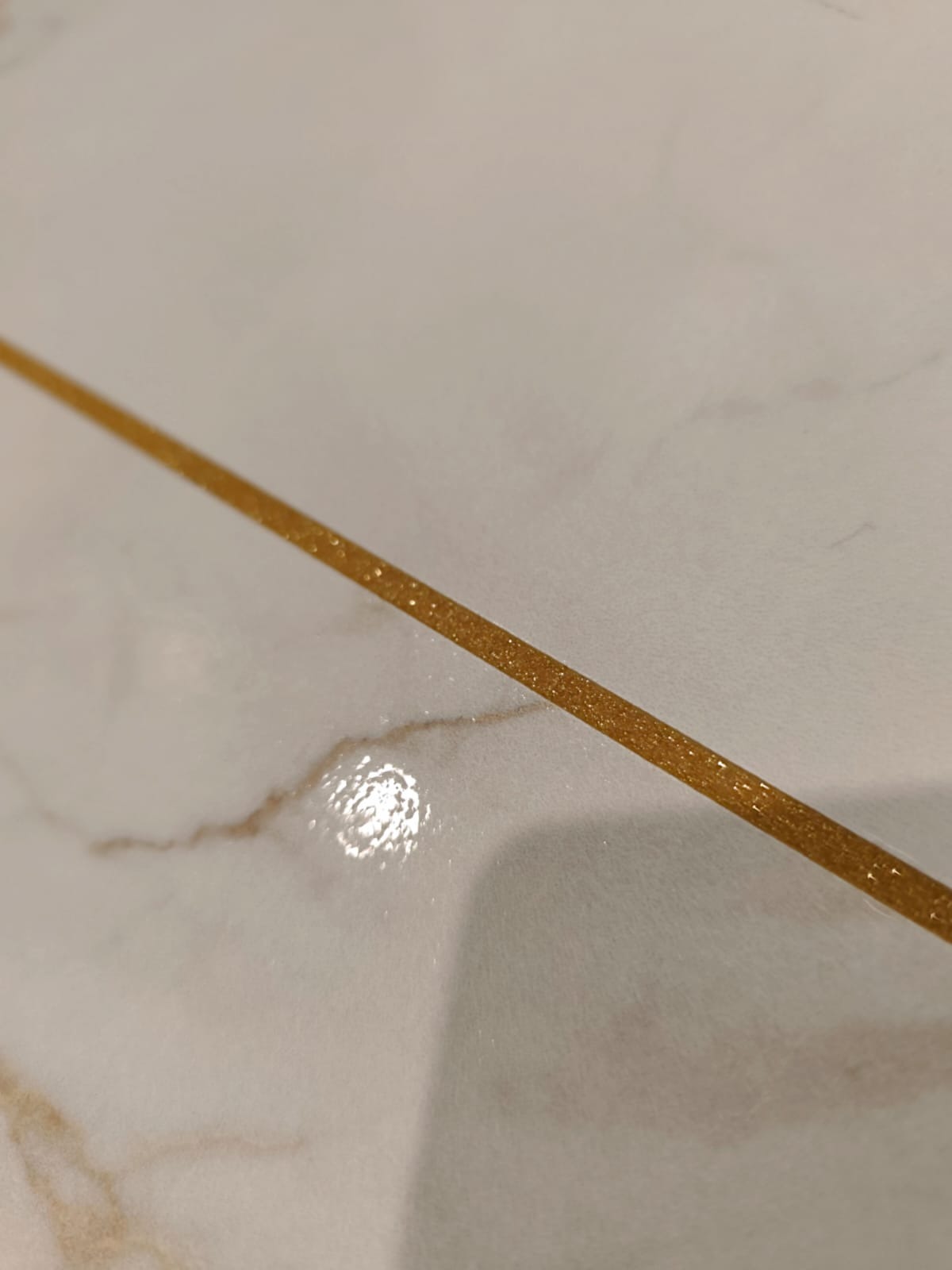 Błyszcząca, złota fuga na białych, marmurkowych płytkach podłogowych, ułożonych ukośnie. Detal wykończenia z bliska.