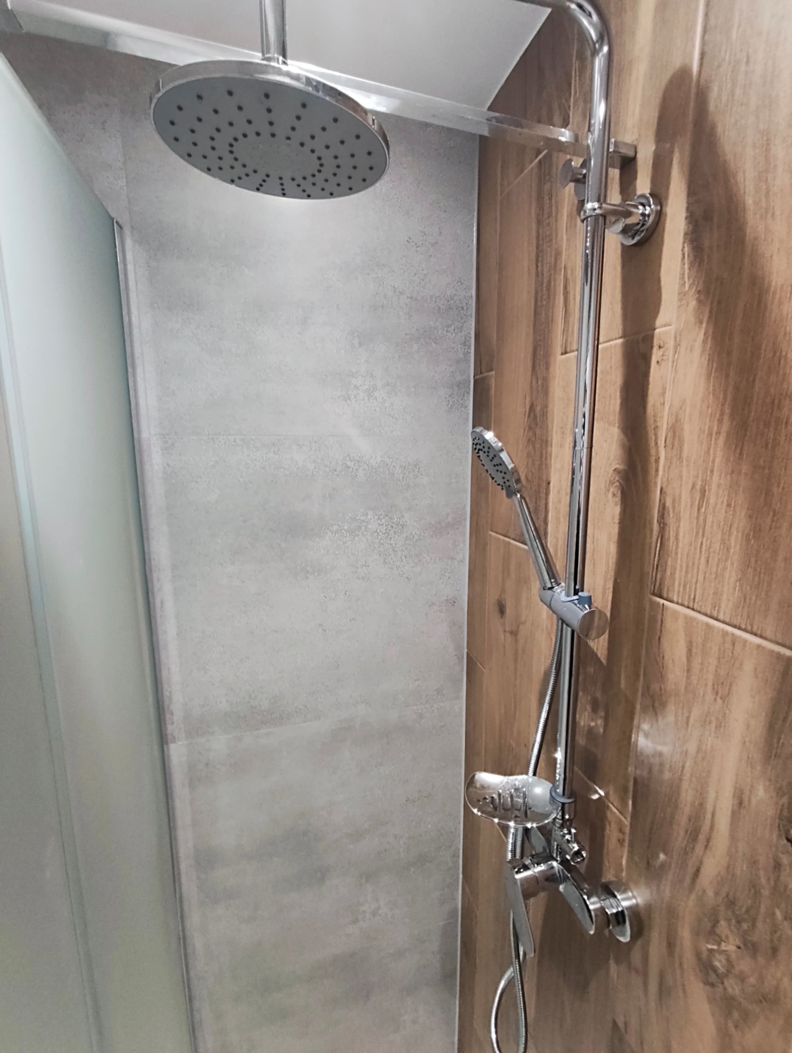 Nowoczesna kabina prysznicowa z deszczownicą i baterią, wykończona płytkami imitującymi drewno i szarym kamieniem. Minimalistyczny design, chromowane elementy.