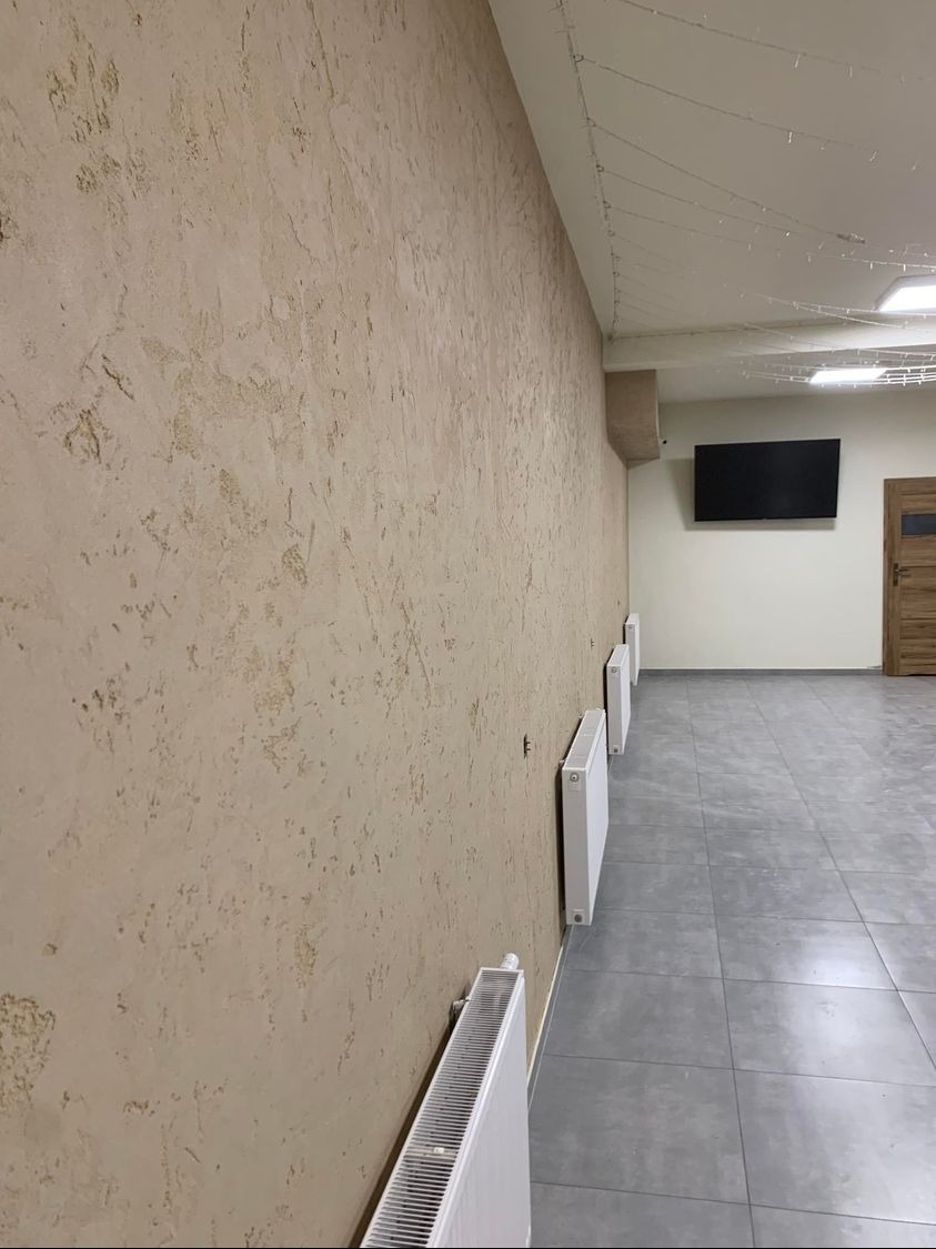 Beżowa ściana z dekoracyjną fakturą, grzejniki, szara podłoga z płytek, telewizor na ścianie w tle. Widok z boku, oświetlenie sufitowe.