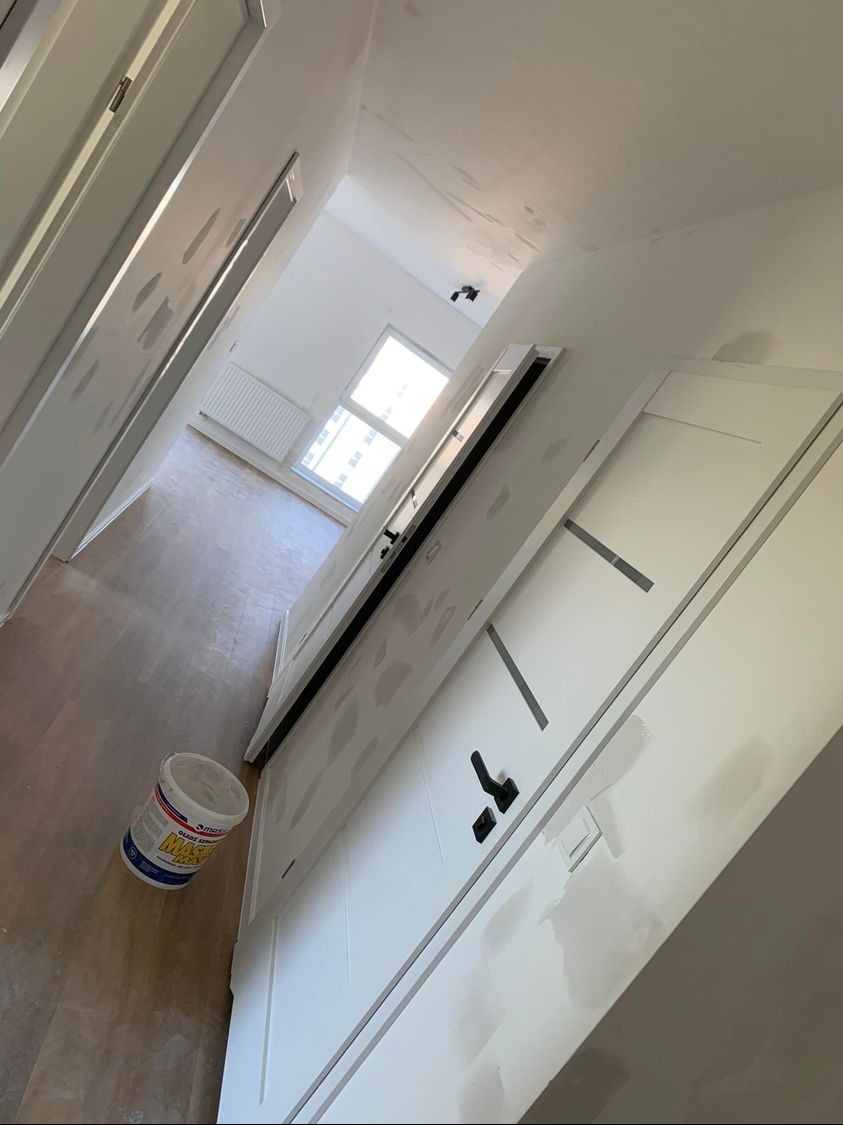 Wnętrze w trakcie prac wykończeniowych: białe ściany z widocznymi śladami po gładzi, drewnopodobna podłoga, białe drzwi z czarną klamką i wiadro z masą szpachlową.