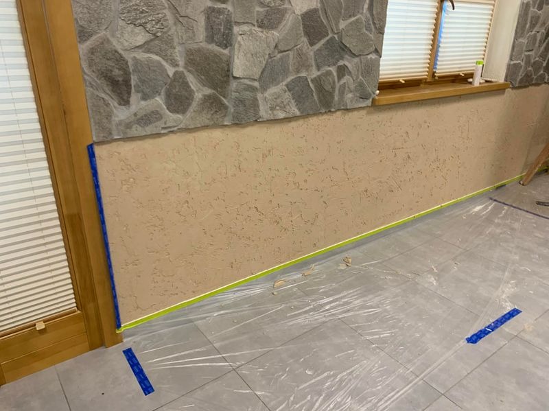 Ściana z nałożoną strukturą, zabezpieczona taśmą malarską i folią ochronną na podłodze, w trakcie prac wykończeniowych. Fragment kamiennej ściany u góry.