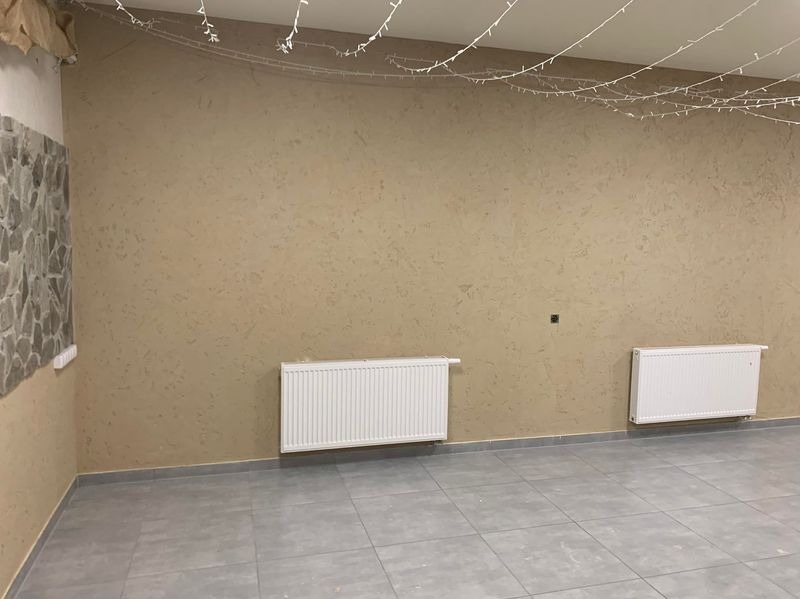 Beżowa ściana z dekoracyjną fakturą, dwa białe grzejniki, szara podłoga z płytek i girlanda świetlna pod sufitem. Fragment ściany wykończony kamieniem dekoracyjnym.