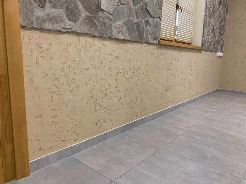 Ściana z dekoracyjną, beżową strukturą tynku, szara podłoga z płytek, kamienna okładzina ściany i drewniana rama okienna z żaluzjami w jasnym wnętrzu.