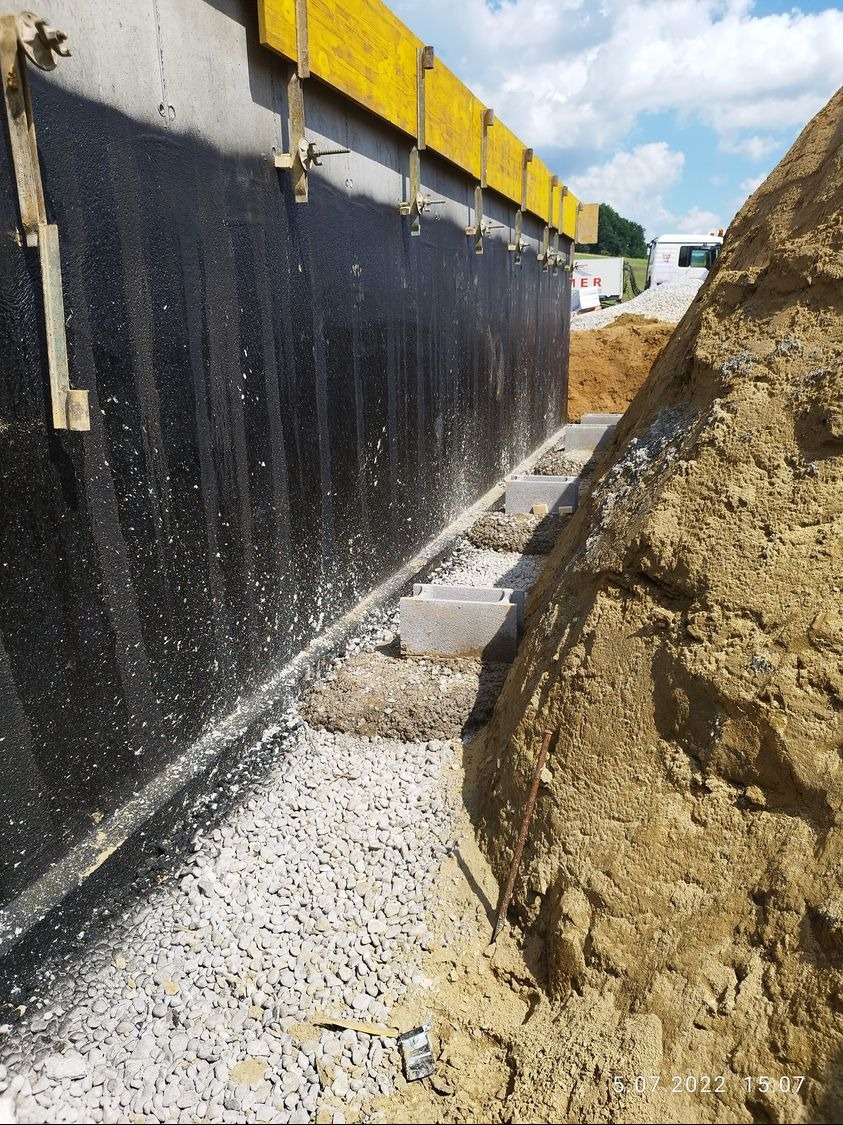 Izolacja pionowa fundamentu z deskowaniem i drenażem. Widoczne bloczki betonowe i ziemia. Prace ziemne w toku, przygotowanie do dalszych etapów budowy.