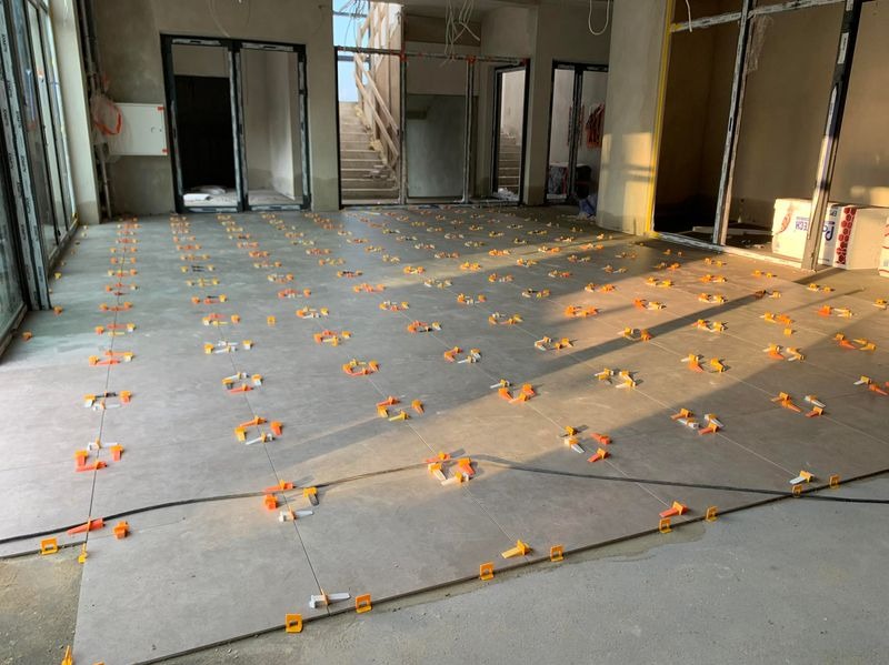 Układanie szarych płytek podłogowych z pomarańczowymi klipsami poziomującymi w przestronnym wnętrzu z oknami i schodami w tle. Etap prac wykończeniowych.