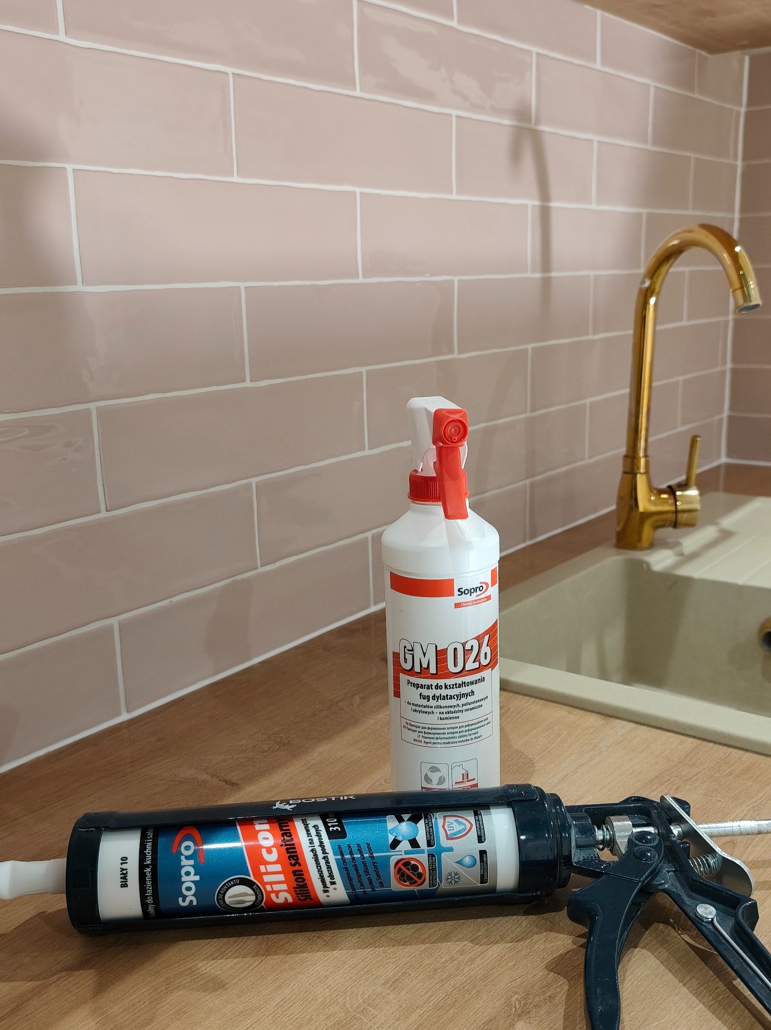 Świeżo położone, różowe płytki w łazience z złotą baterią umywalkową, preparatem do fug i silikonem sanitarnym marki Sopro na blacie.