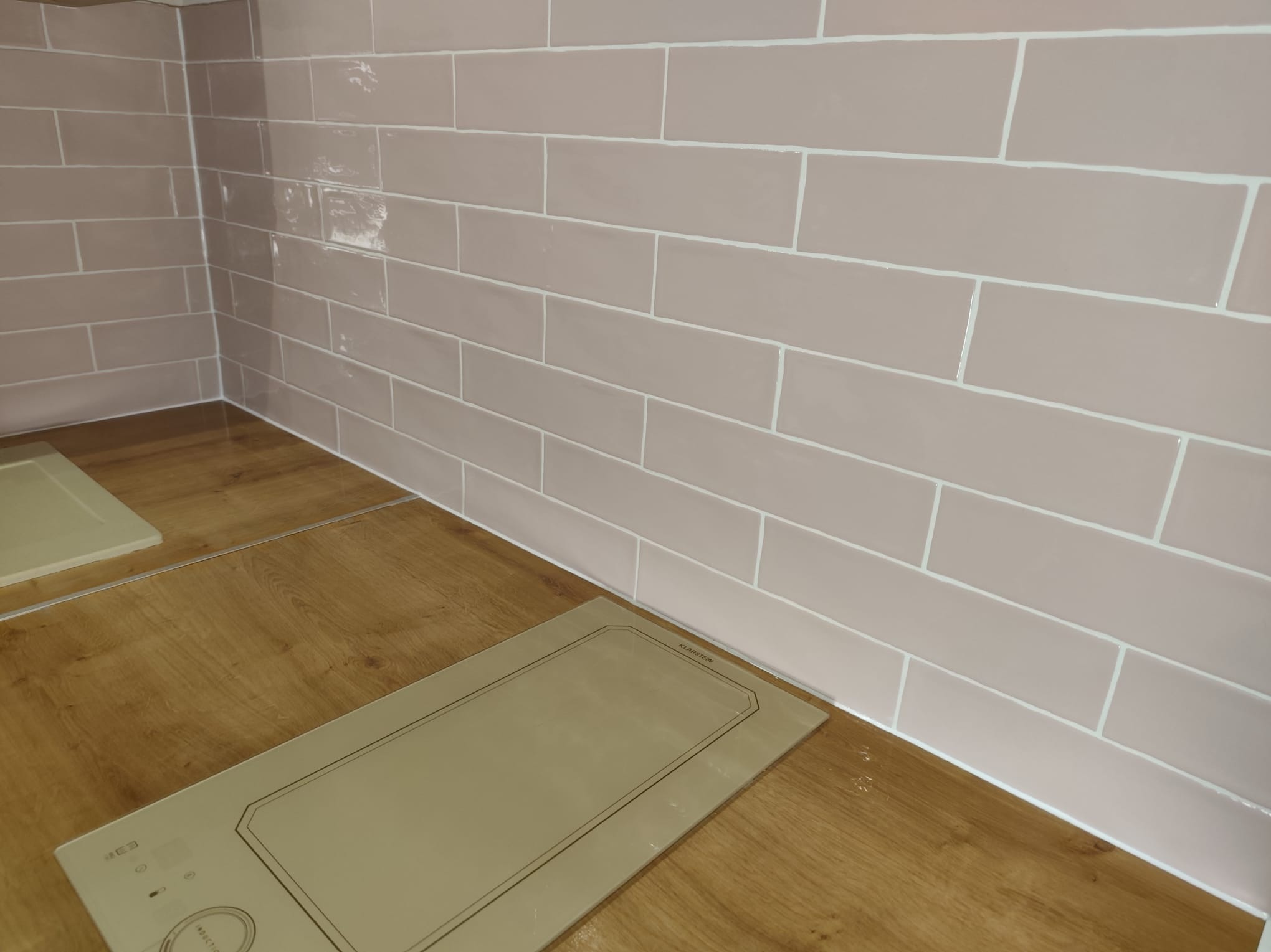 Ściana wyłożona podłużnymi, beżowymi płytkami z białymi fugami, przylegająca do drewnianego blatu z płytą indukcyjną. Widoczne niedociągnięcia w fugowaniu.