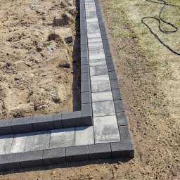 Kompleksowe Wykończenia Wnętrz Andrzej Berhorst - Układanie obrzeży z kostki brukowej w ogrodzie, widok z góry. Częściowo wypełnione piaskiem, w tle taczka i krzesła ogrodowe. Słoneczny dzień.