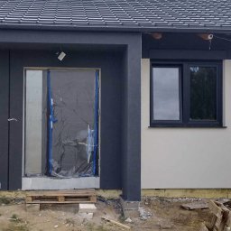 Kompleksowe Wykończenia Wnętrz Andrzej Berhorst - Fragment elewacji domu w trakcie prac wykończeniowych: szara stolarka okienna i drzwiowa, częściowo otynkowana ściana w kolorze złamanej bieli, zabezpieczone drzwi.