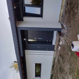 Kompleksowe Wykończenia Wnętrz Andrzej Berhorst - Nowoczesny dom jednorodzinny z szarą stolarką okienną i drzwiową, białą elewacją oraz fragmentem ocieplenia widocznym przy gruncie. Widoczna boczna elewacja budynku.