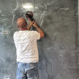 Kompleksowe Wykończenia Wnętrz Andrzej Berhorst - Pracownik szlifuje ścianę z dekoracyjną szarą strukturą przypominającą marmur, używając szlifierki. Widoczne gniazdka elektryczne i klimatyzator.