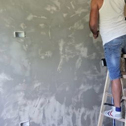 Kompleksowe Wykończenia Wnętrz Andrzej Berhorst - Pracownik na drabinie aplikuje dekoracyjną, szarą farbę na ścianę w pomieszczeniu. Widoczne puszki elektryczne i zabezpieczona taśmą listwa sufitowa.