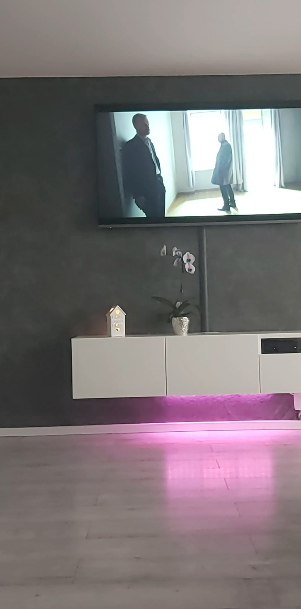 Szara ściana z efektem dekoracyjnym, biała szafka RTV z podświetleniem LED, telewizor z filmem. Minimalistyczny design, nowoczesne wykończenie wnętrz.