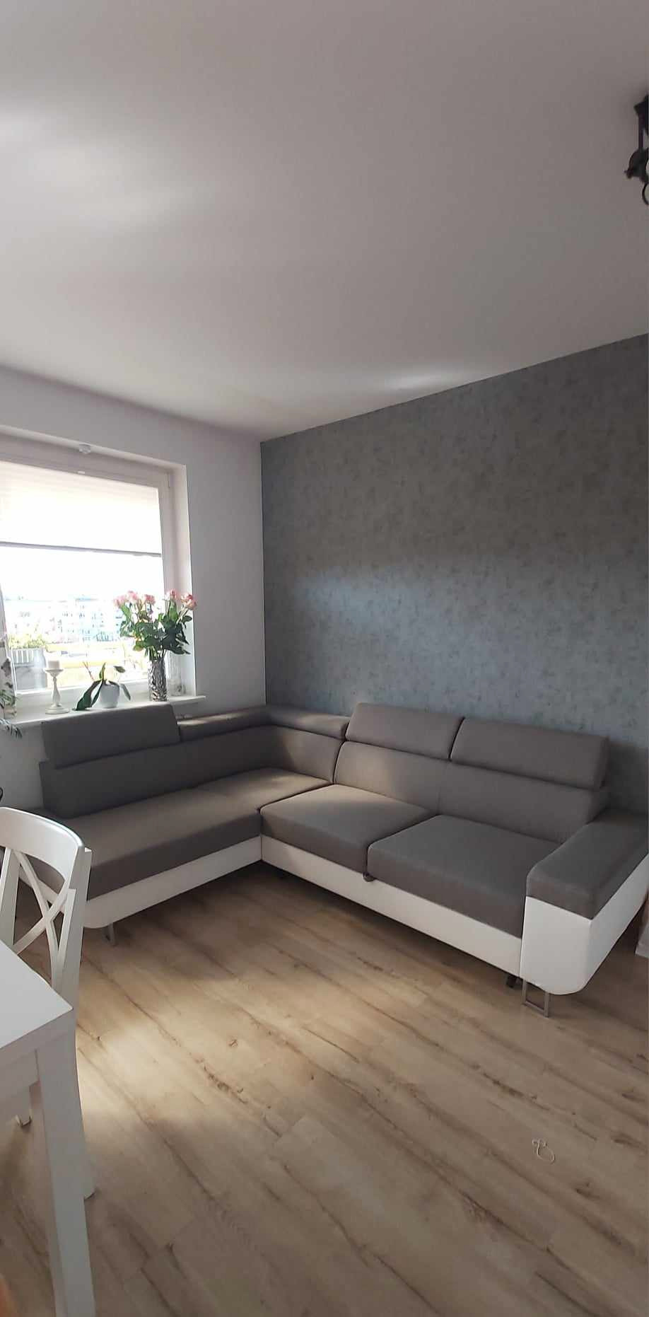 Szary narożnik z białymi bokami w salonie po wykończeniu w Gdańsku. Na ścianie dekoracyjna szara farba, jasna podłoga, okno z kwiatami.