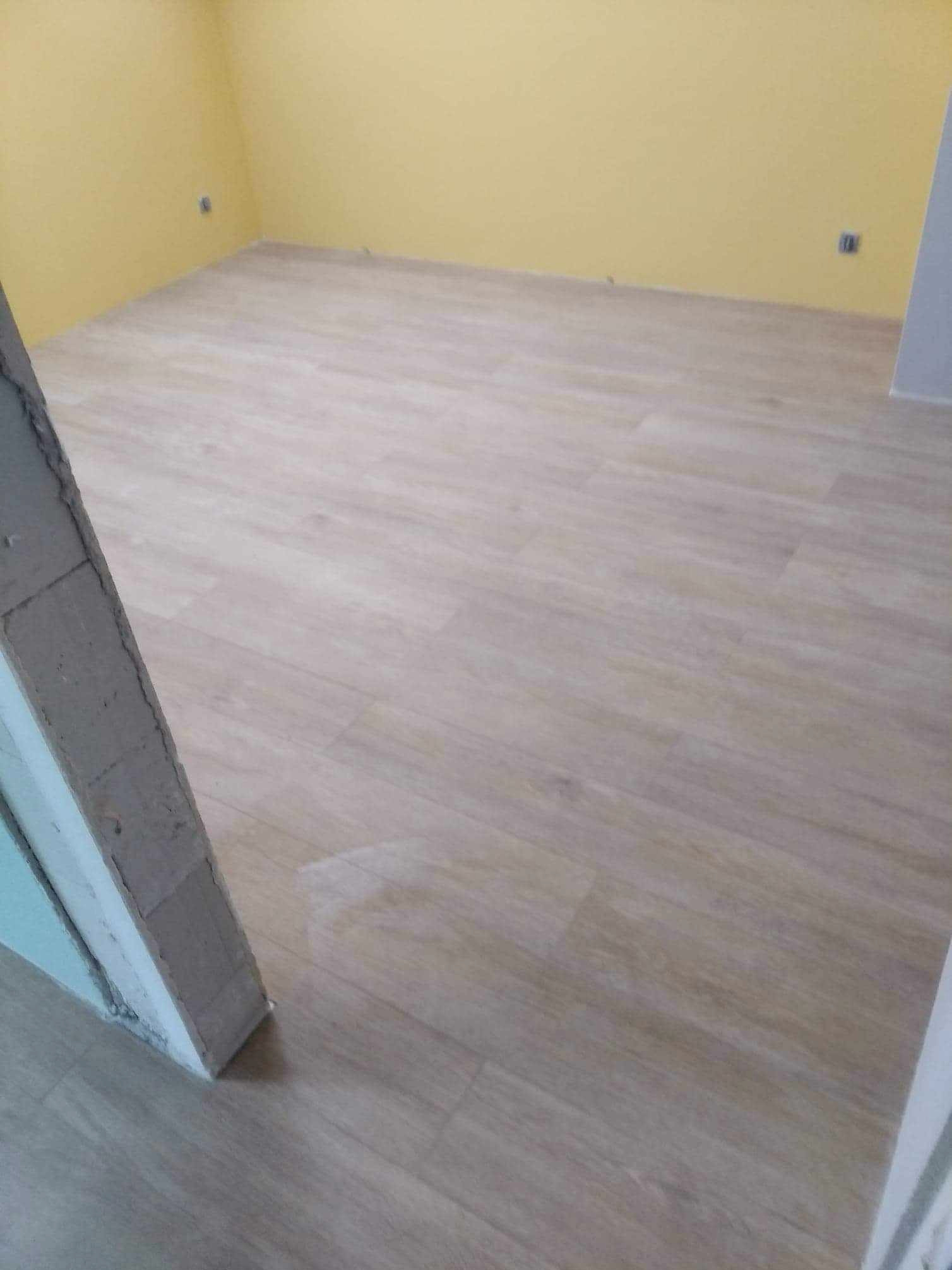 Jasne panele podłogowe ułożone w pustym pokoju z żółtymi ścianami. Widoczna ościeżnica drzwi po lewej stronie kadru. Minimalistyczne wykończenie wnętrza.