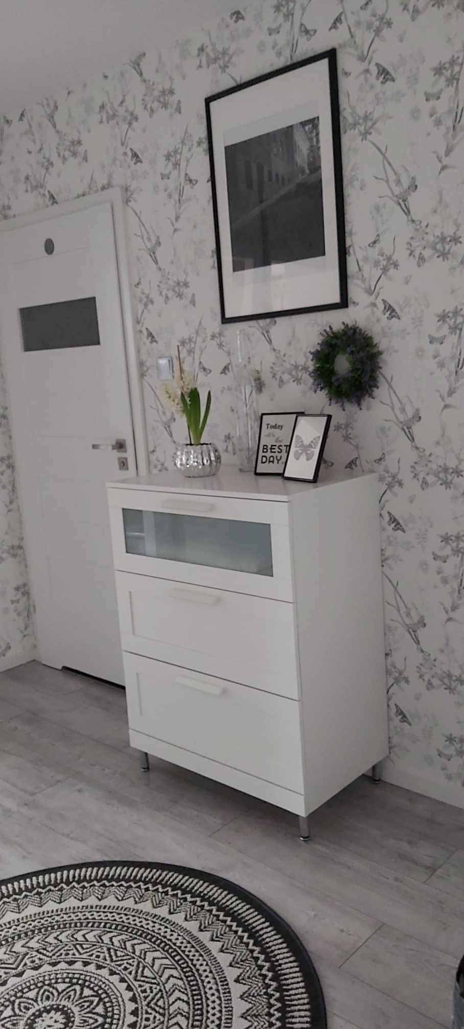 Biała komoda z trzema szufladami, dekoracje na blacie, drzwi i wzorzysta tapeta w tle. Element wykończenia wnętrza w odcieniach szarości i bieli.