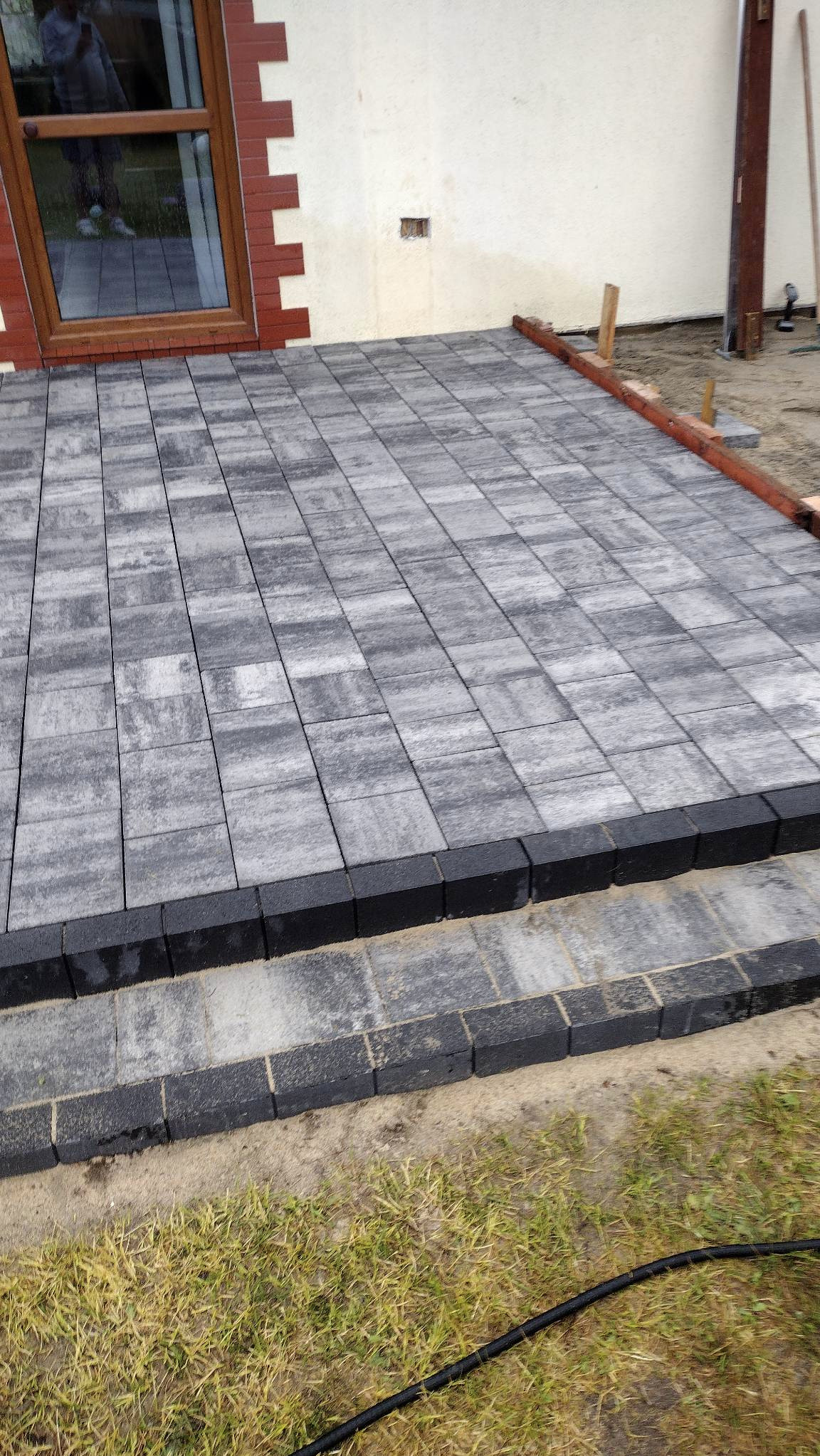 Ułożona kostka brukowa w odcieniach szarości tworząca taras przy budynku z oknem. Widoczne obrzeża i fragment trawnika. Ukończony etap prac.