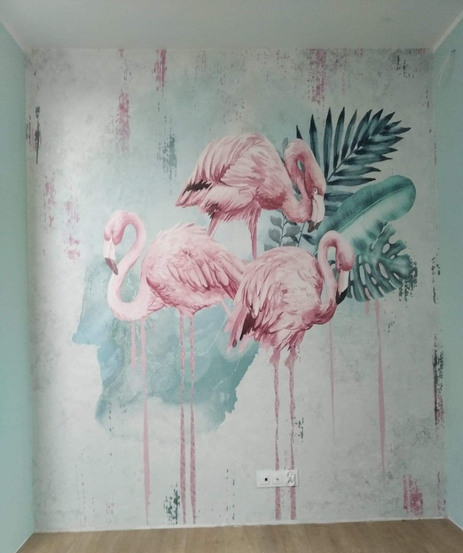 Wnętrze pokoju z fototapetą przedstawiającą trzy różowe flamingi i liście na tle w odcieniach błękitu i różu. Podłoga drewniana, ściana boczna w kolorze miętowym.
